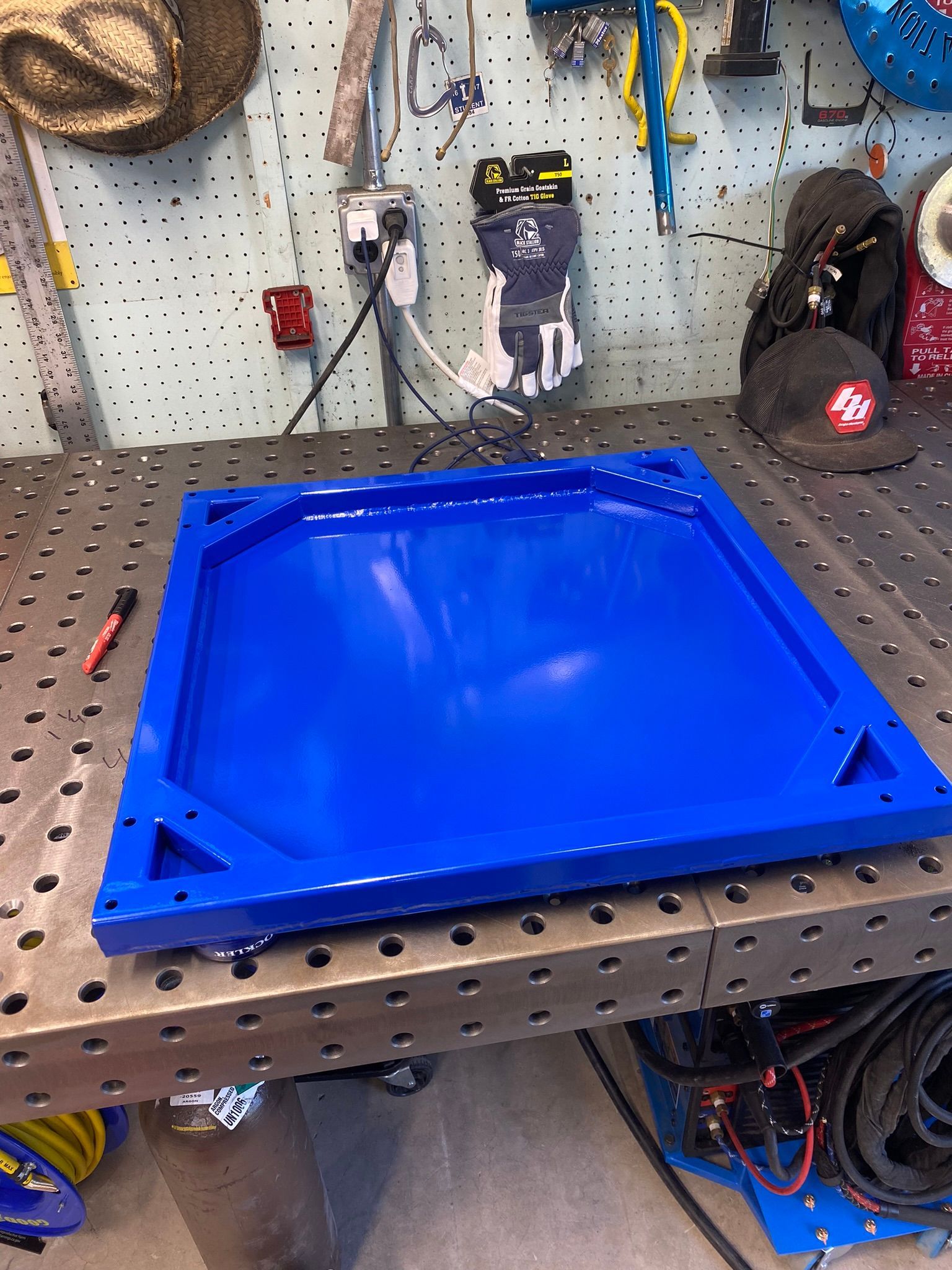 A bright blue metal tray with triangular corner cutouts sitting on a perforated welding table in a workshop.
