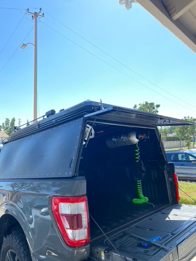 A grey pickup truck with a black metal bed cap featuring an open rear hatch and a neon green coil cable hanging inside.