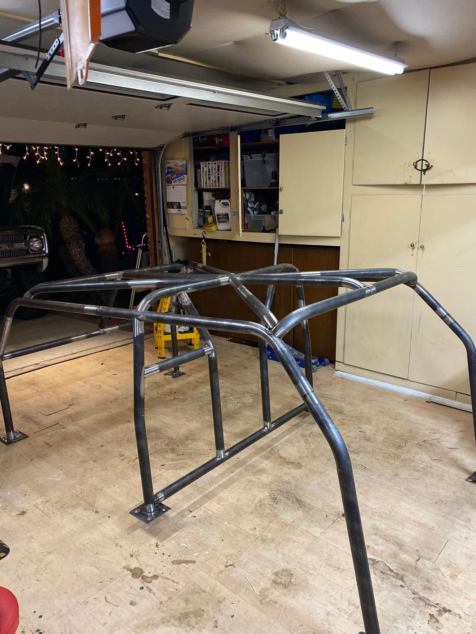 A metal roll cage frame sits in a cluttered garage, resting on the concrete floor under bright overhead lights.