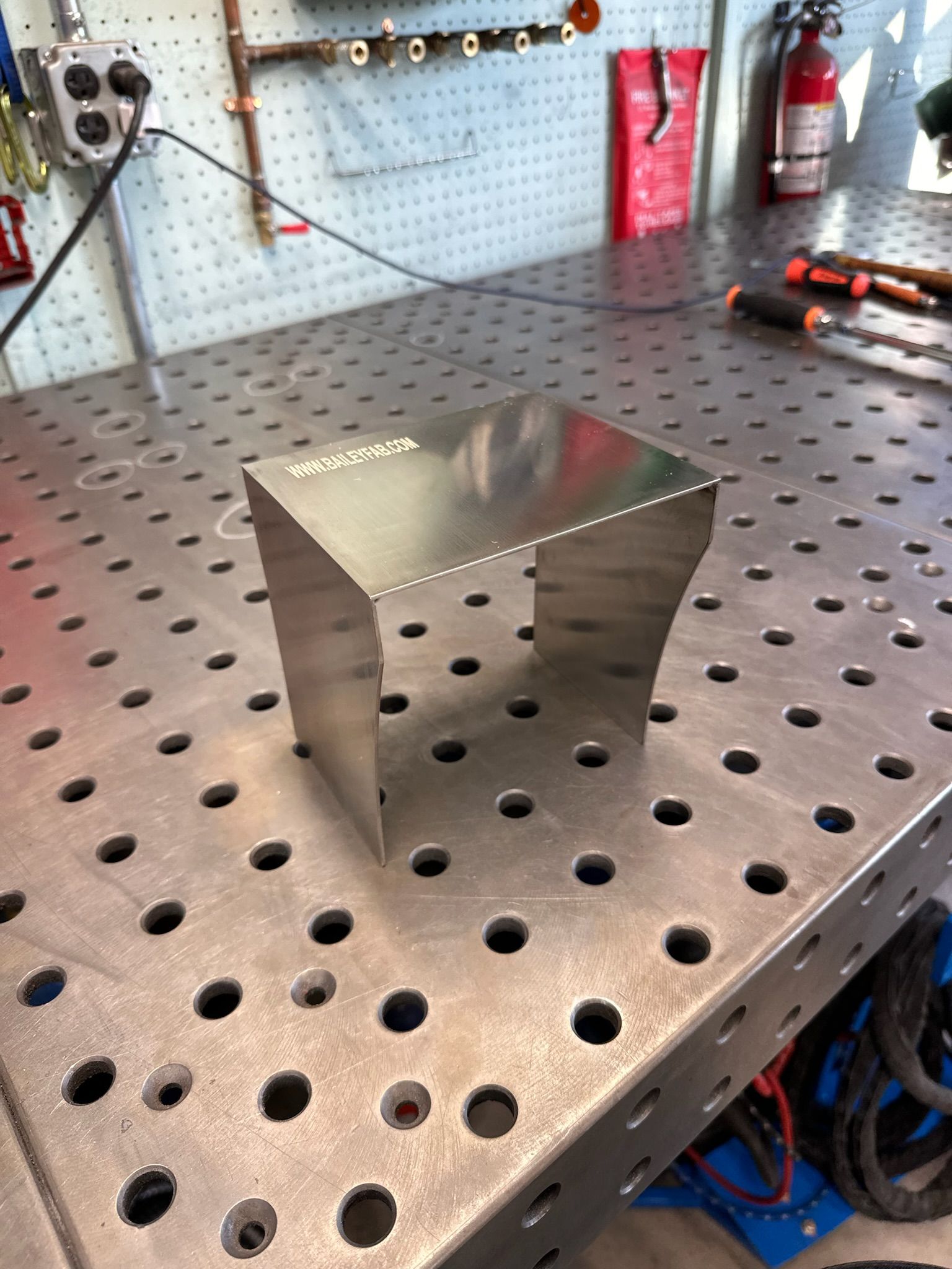 A U-shaped metal bracket sits on a perforated metal welding table in a workshop.