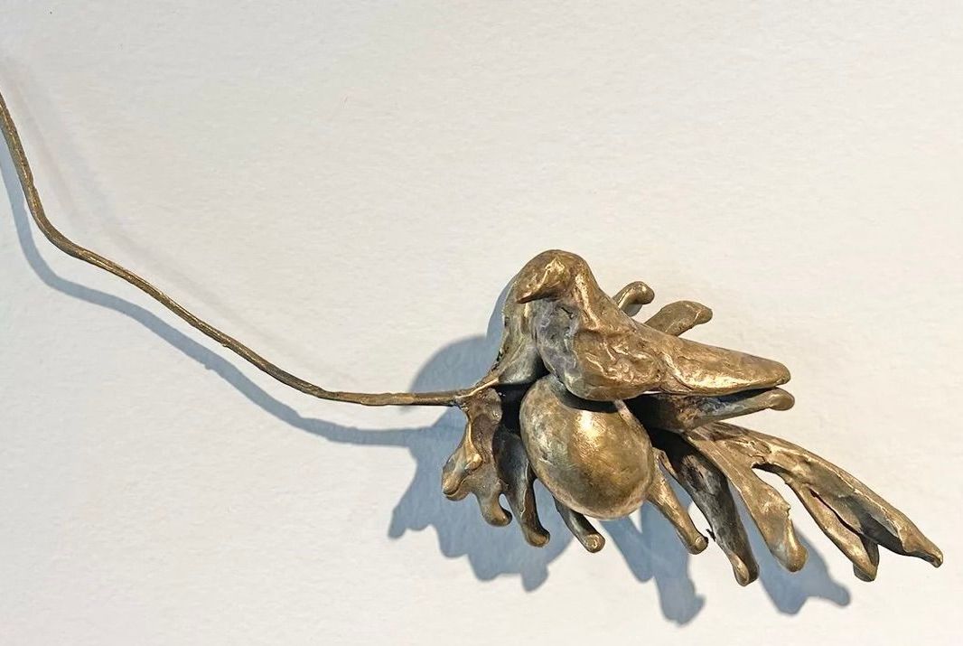 A metallic, bronze-colored sculpture with organic, jagged shapes hanging from a thin, textured chain against a white wall.