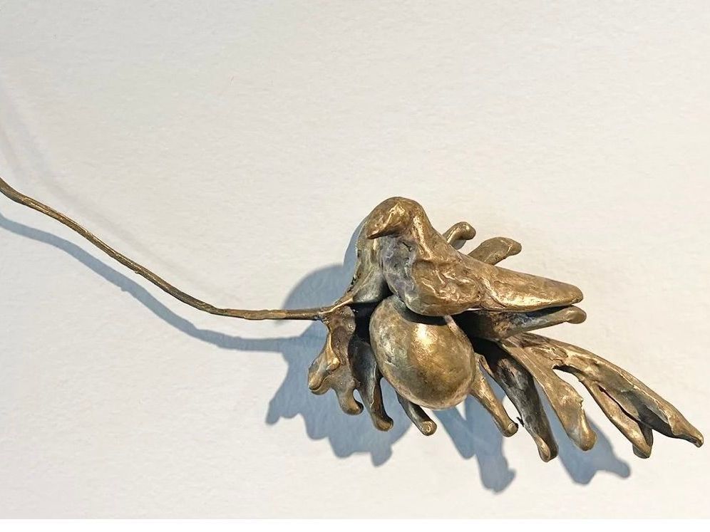 A brass sculpture of a bird perched on leafy branches, extended on a thin wire against a plain white background.