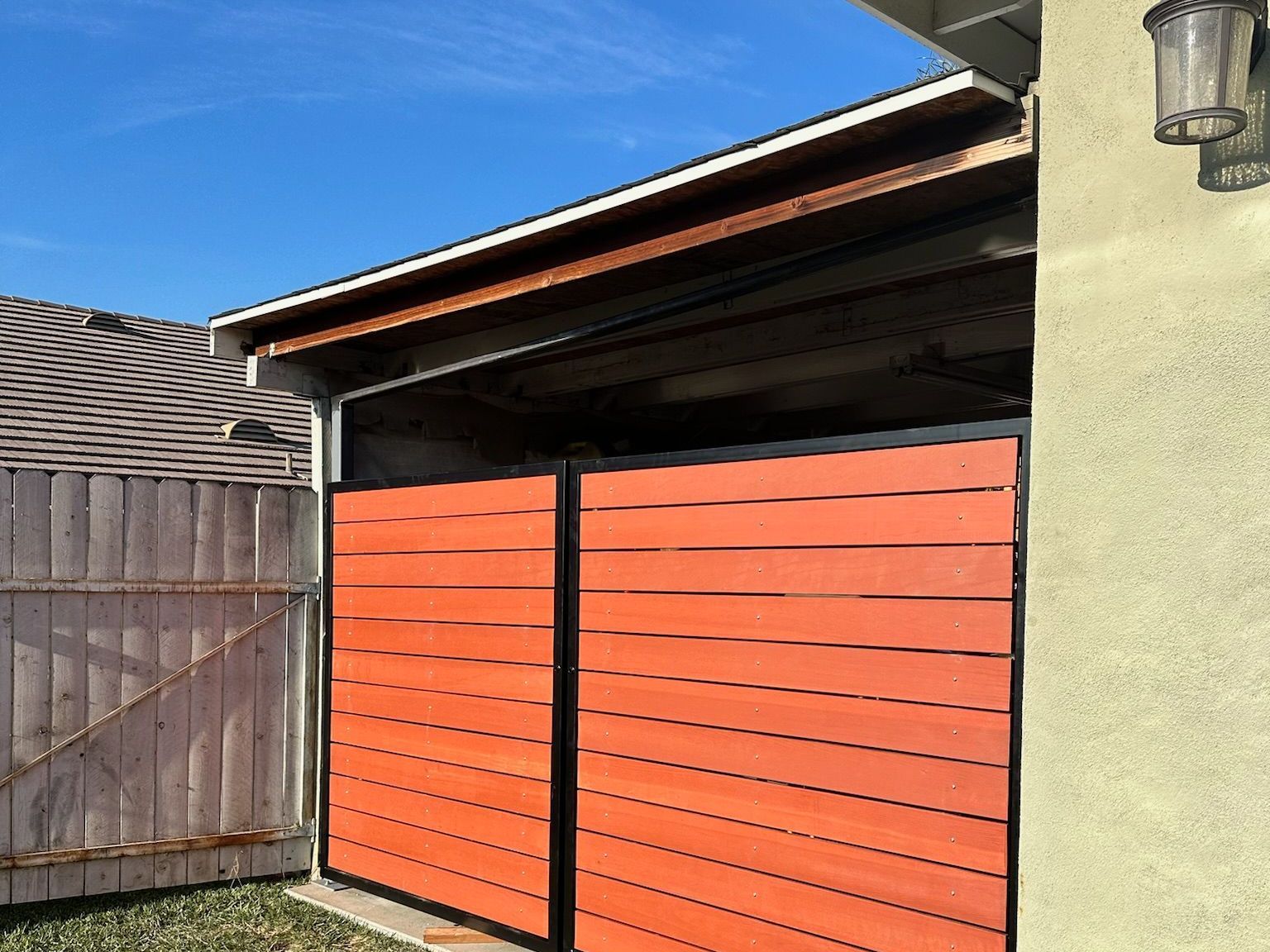 A horizontal orange wooden gate stands in front of a garage, next to a wood fence under a blue sky.