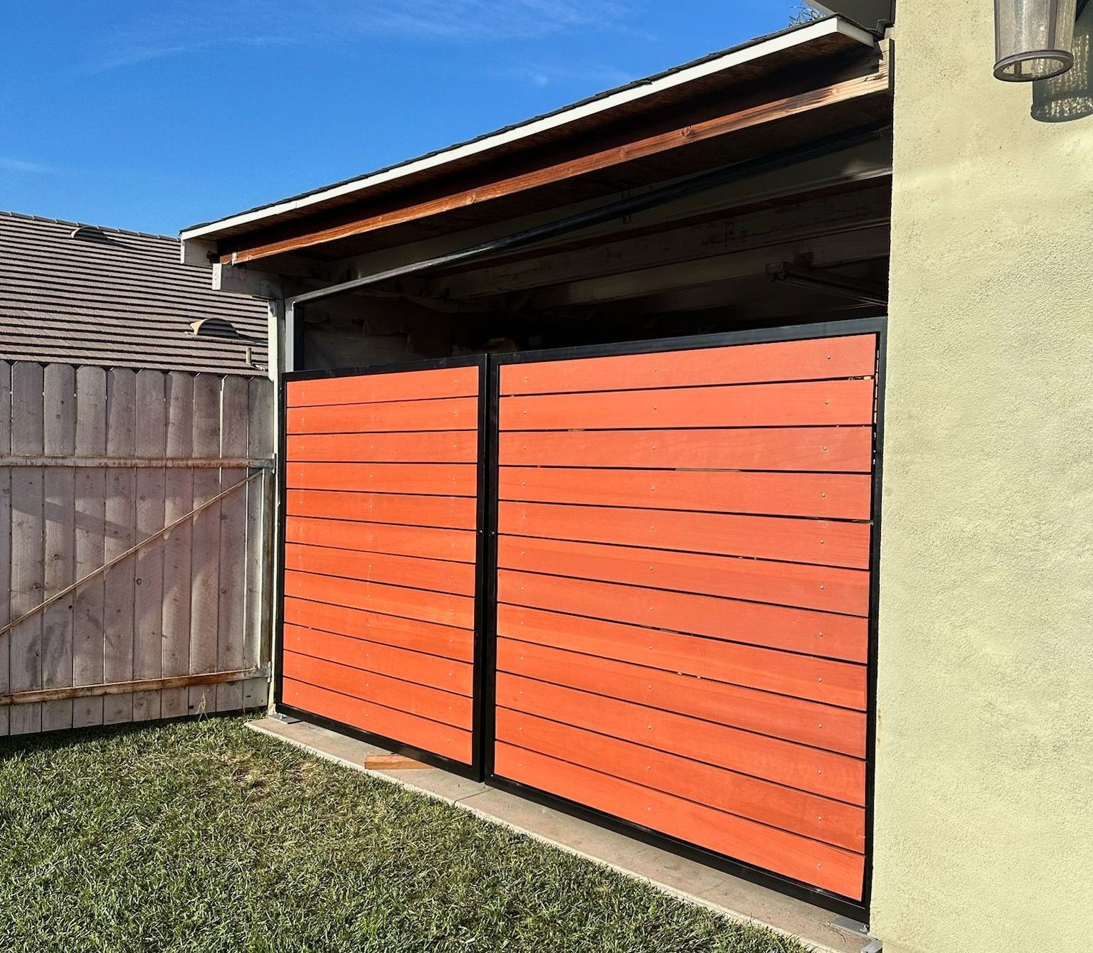 Two modern metal-framed gates with horizontal orange wooden slats installed in a residential driveway.