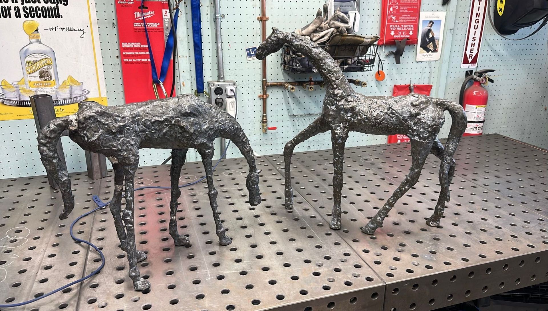 Two welded metal sculptures of four-legged animals stand on a perforated metal workbench in a workshop.