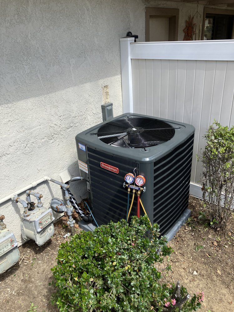 HVAC unit outside a building, with gauges attached. Gray and black unit near bushes and a white fence.
