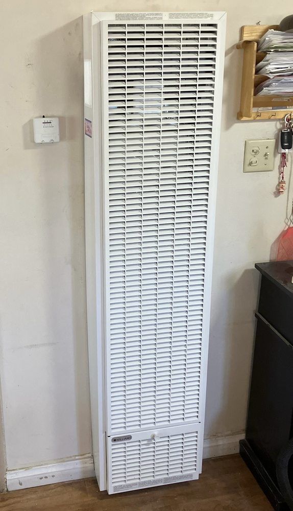 White wall-mounted gas heater with a rectangular vent cover, next to a wall-mounted thermostat, in an indoor setting.
