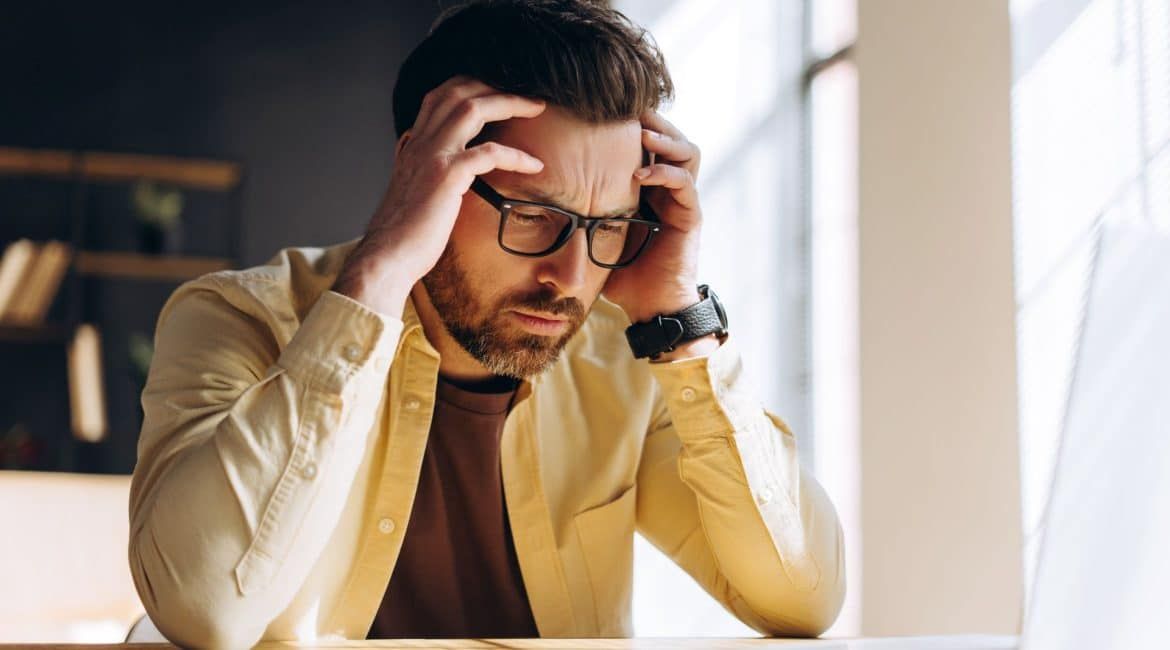Man in glasses and yellow shirt holding his head at a desk, looking stressed or frustrated.