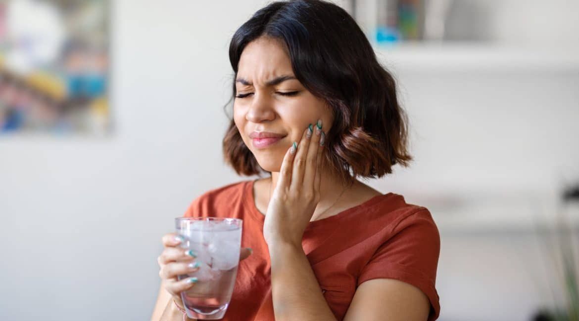 Woman holding her cheek and grimacing while holding a glass of water, suggesting tooth pain.