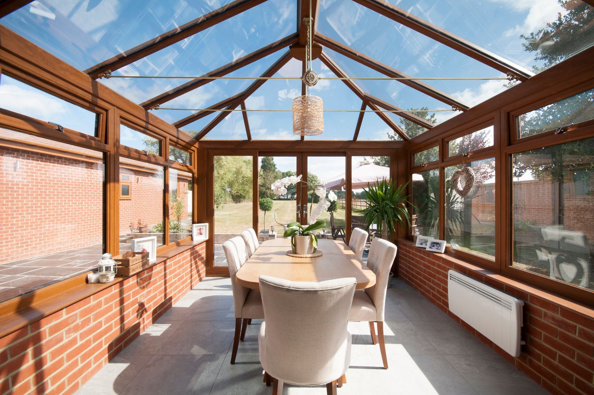 Sunroom with glass roof and windows, wooden frames, brick walls, and dining table set for guests.