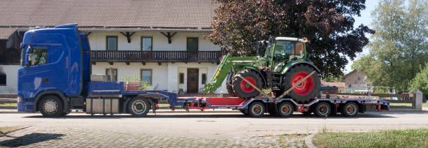 A blue truck is carrying a tractor on a trailer.