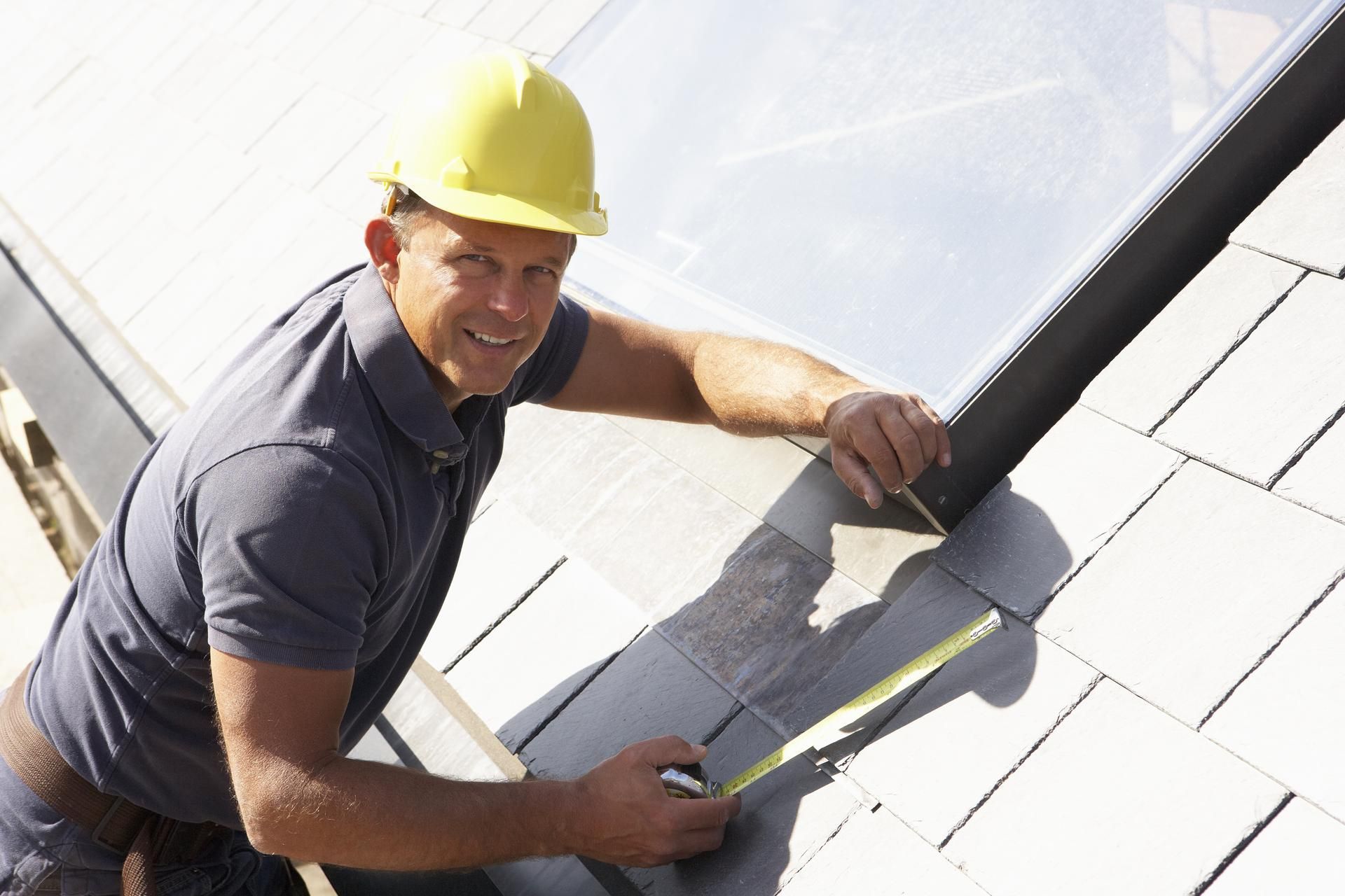 Roofer in a hard hat smiles while measuring tiles on a roof, near a skylight.