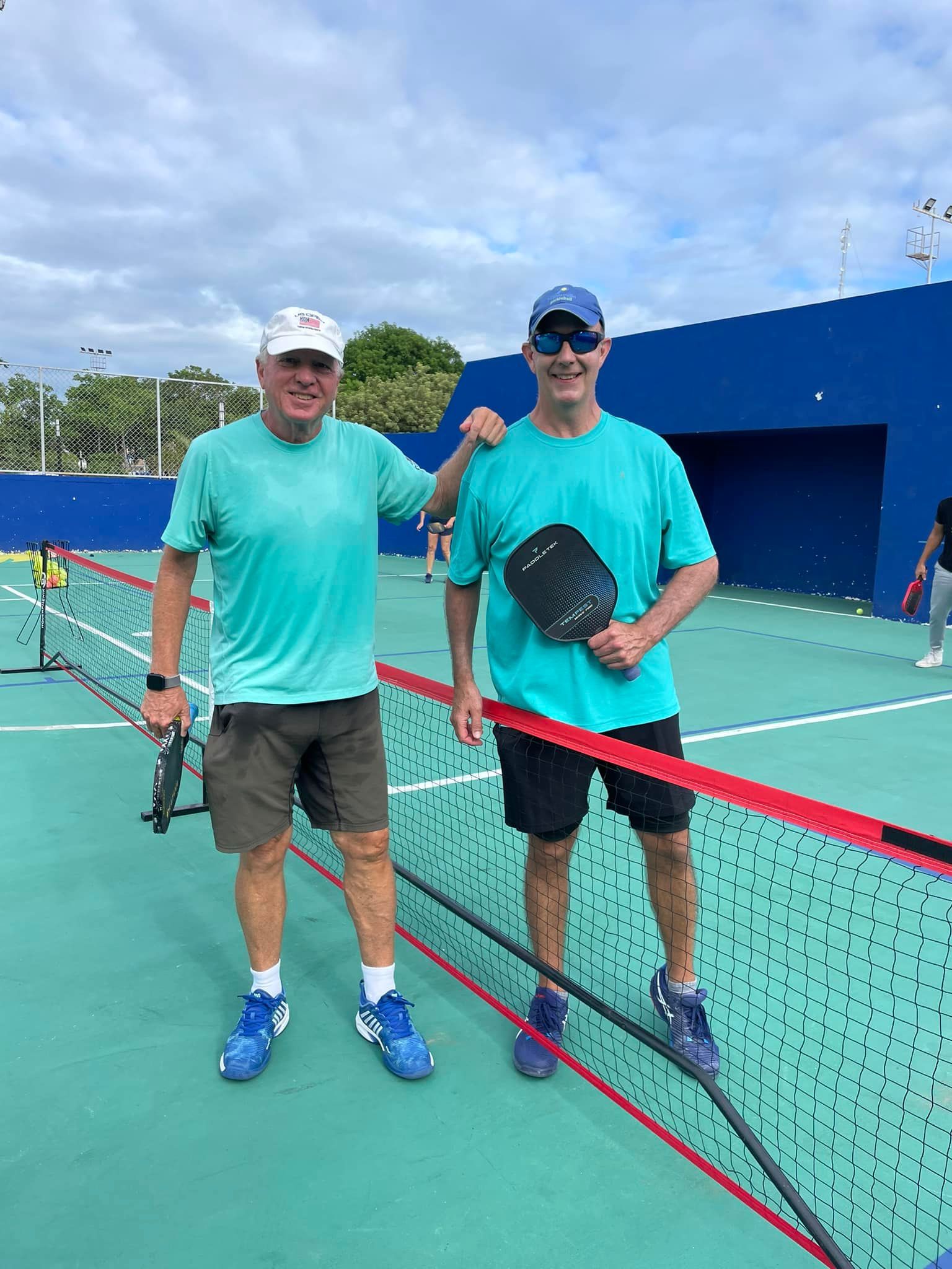 Two men are standing next to each other on a pickleball court at the Playa Del Carmen Pickleball Club in Playa Del Carmen, Mexico 