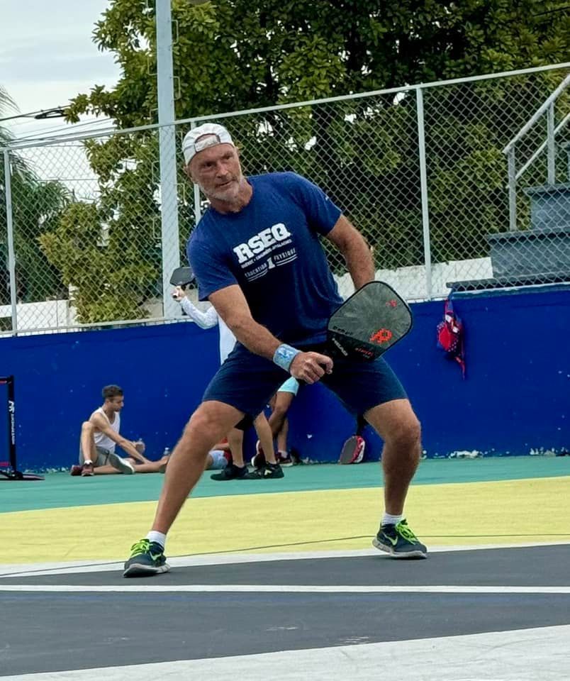 A man in a blue shirt that says rseq is playingpickleball