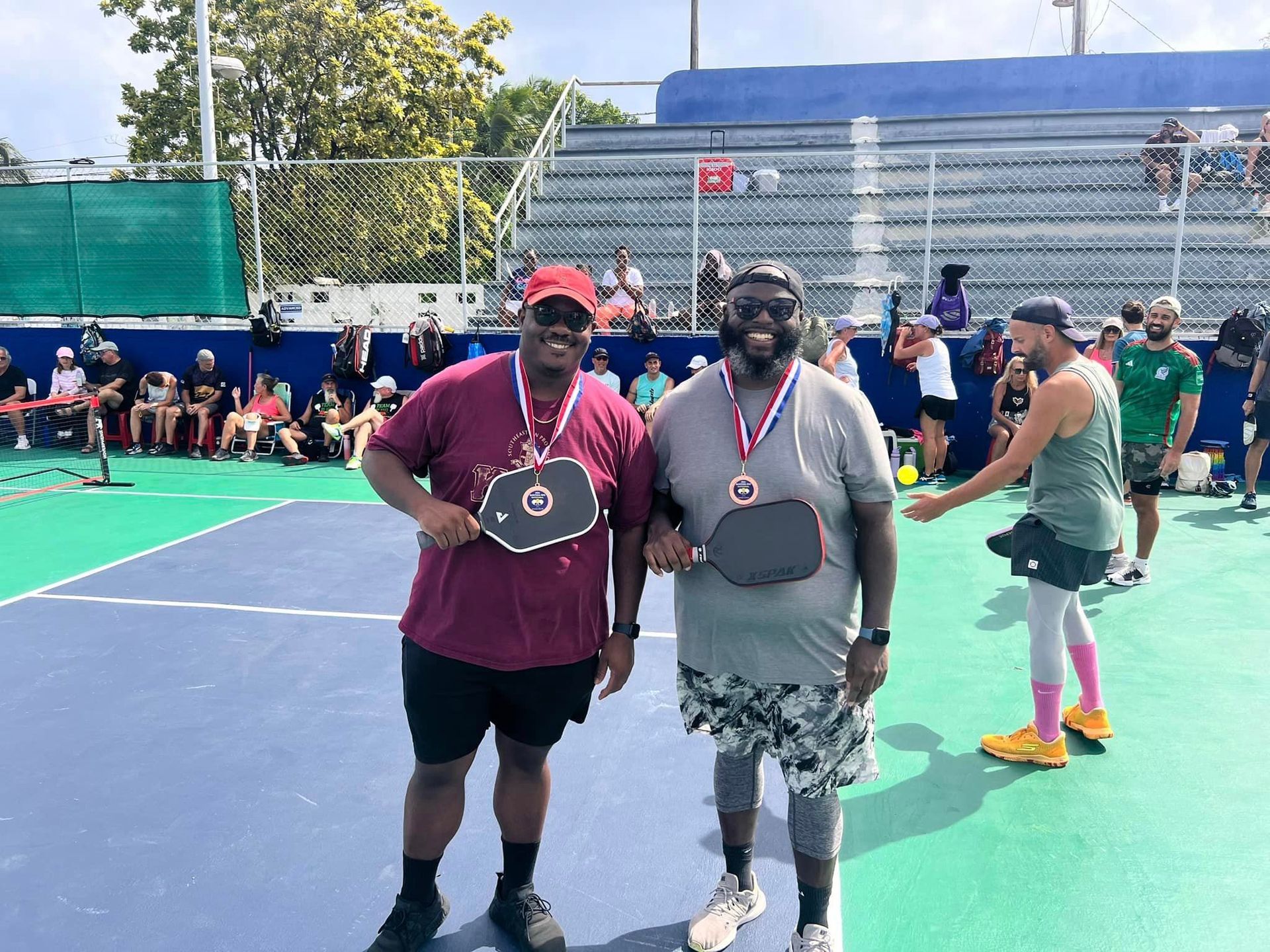 Two men are posing for a picture on a pickleball court. at the Playa Del Carmen Pickleball Club in Playa Del Carmen, Mexico rackets.