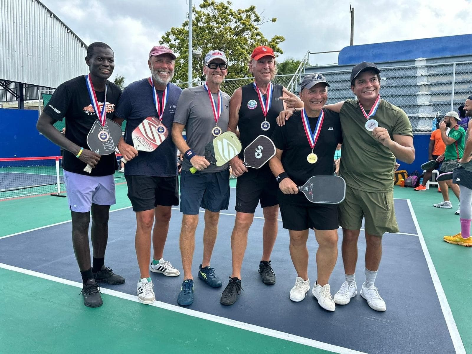 A group of men are posing for a picture on a pickleball court. at the Playa Del Carmen Pickleball Club in Playa Del Carmen, Mexico rackets.