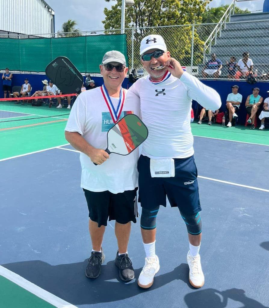 Two men are posing for a picture on a pickleball court at the Playa Del Carmen Pickleball Club in Playa Del Carmen, Mexico rackets.