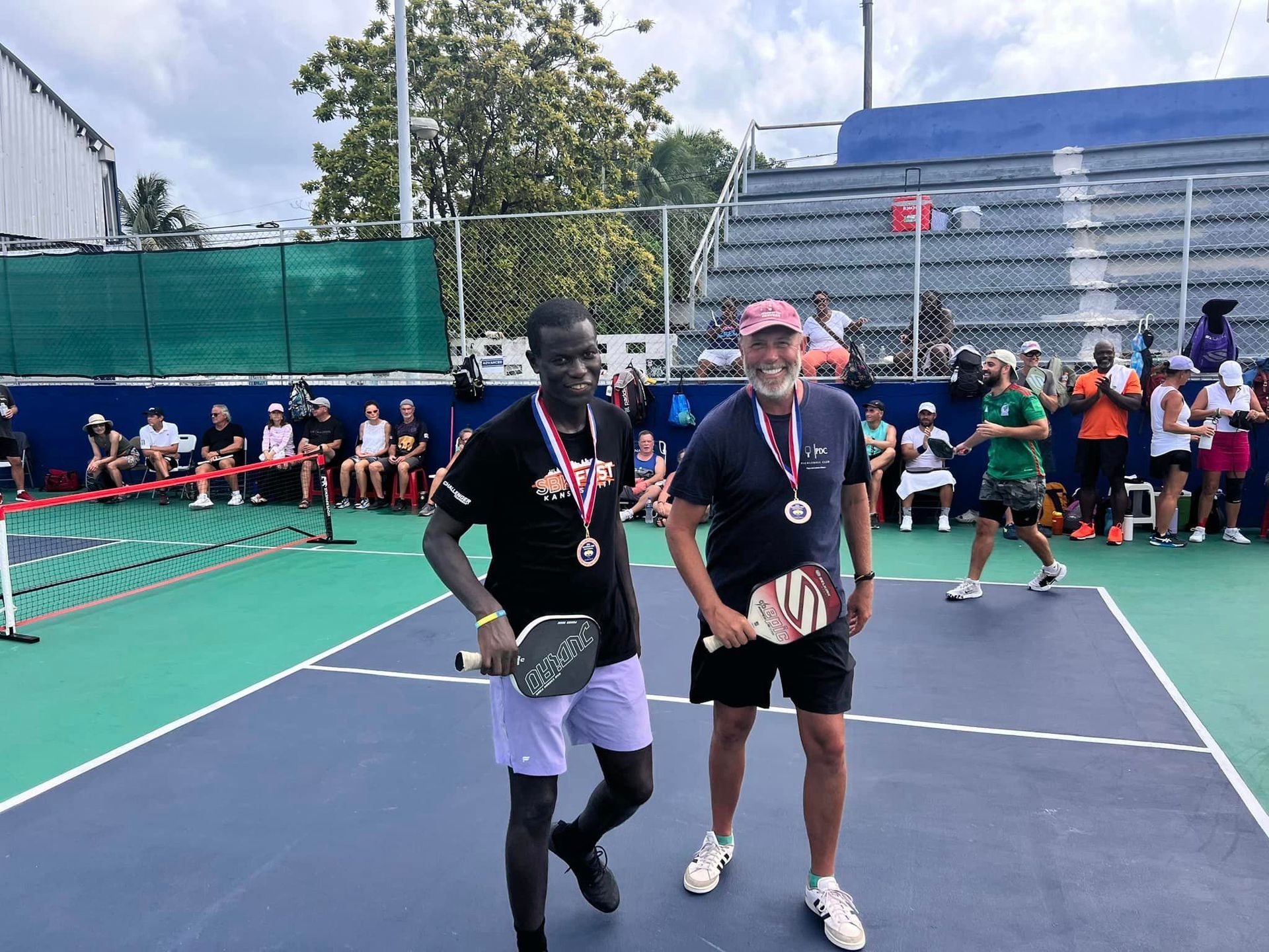Two men are standing on a pickleball court with medals around their necks. at the Playa Del Carmen Pickleball Club in Playa Del Carmen, Mexico rackets.