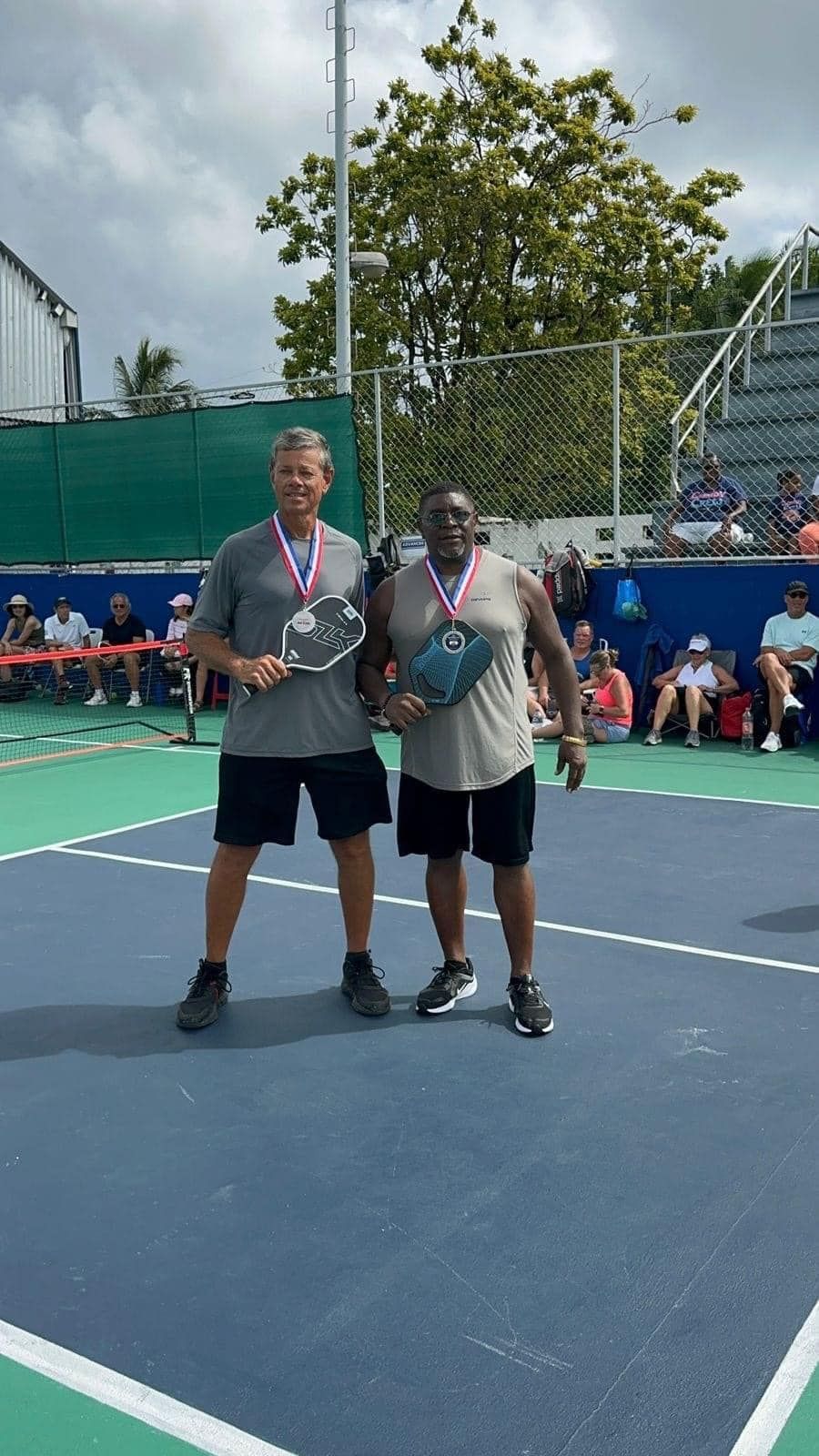Two men are standing on a pickleball court holding pickle ball rackets in at the Playa Del Carmen Pickleball Club in Playa Del Carmen, Mexico rackets.