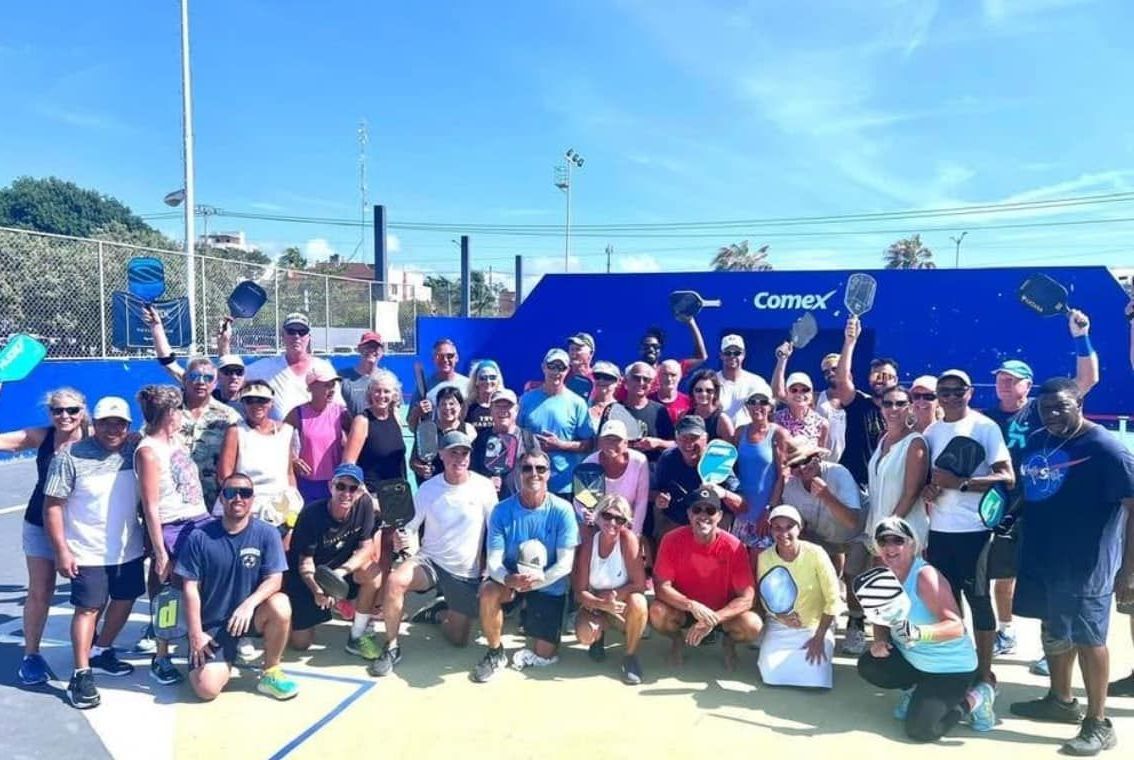 A group of pickleball players are posing for a picture on a pickleball court  at the Playa Del Carmen Pickleball Club in Playa Del Carmen, Mexico rackets.
