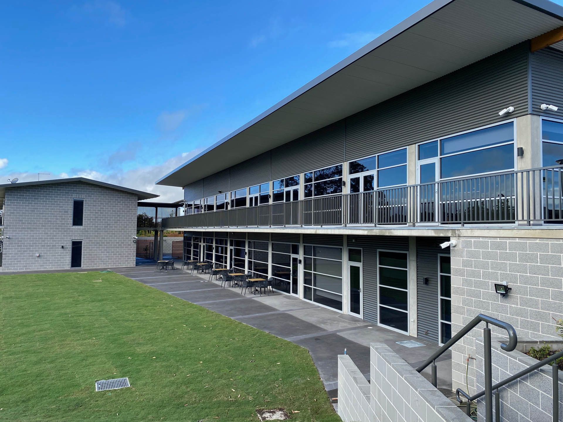Modern two-story building with large windows and a covered walkway, next to a smaller structure on a grassy area under a blue sky— Ray Smith Electrical in Korora, NSW