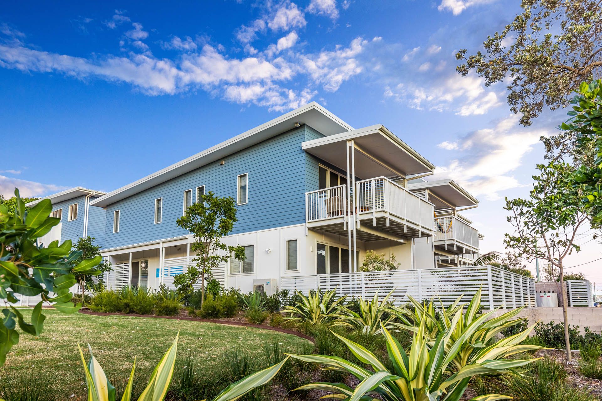 Two-story blue and white building with balconies, under a bright blue sky. Green plants and grass in front— Ray Smith Electrical in Korora, NSW