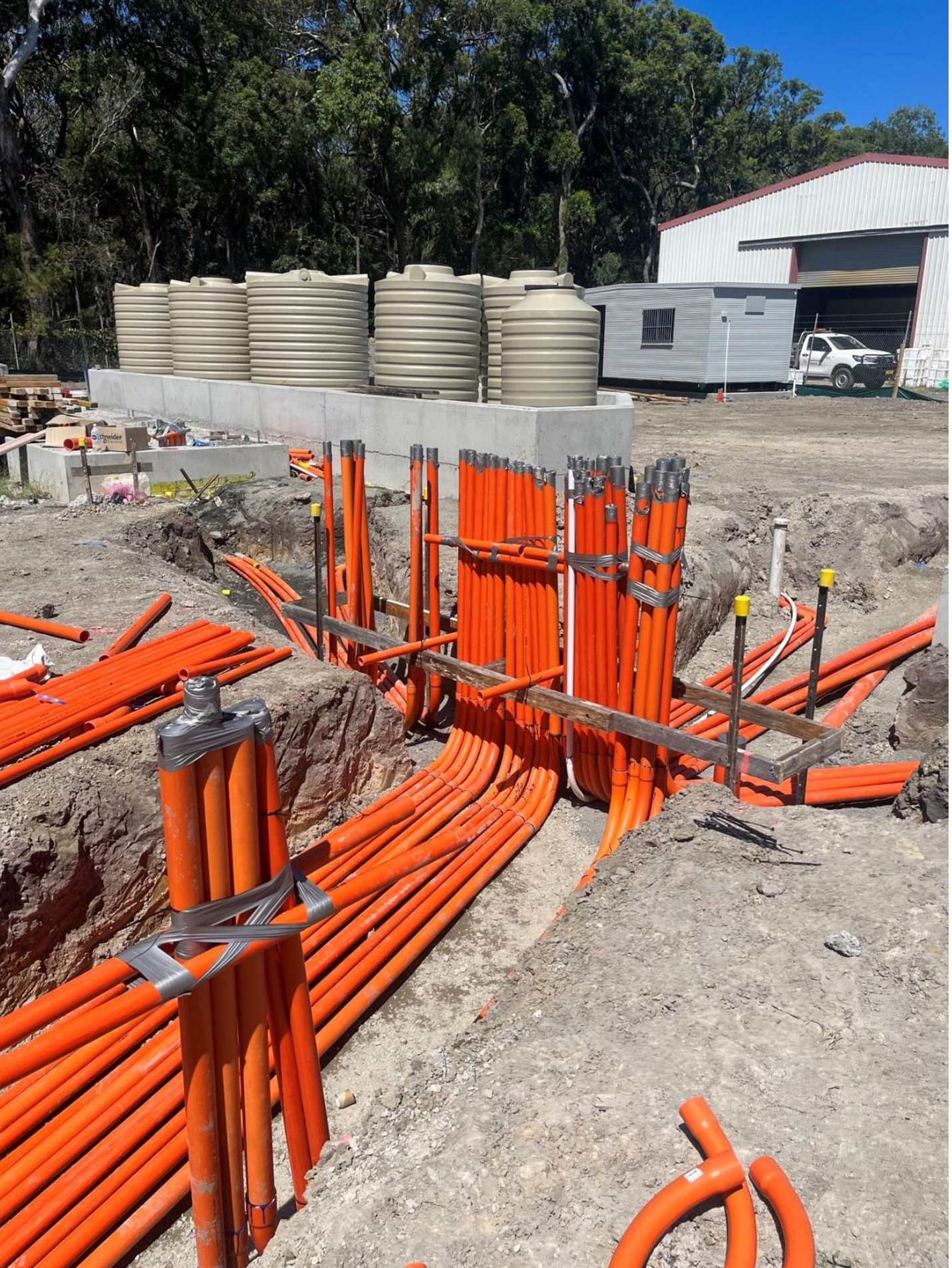 Orange conduit pipes installed in trenches at a construction site, with water tanks and a building visible— Ray Smith Electrical in Korora, NSW