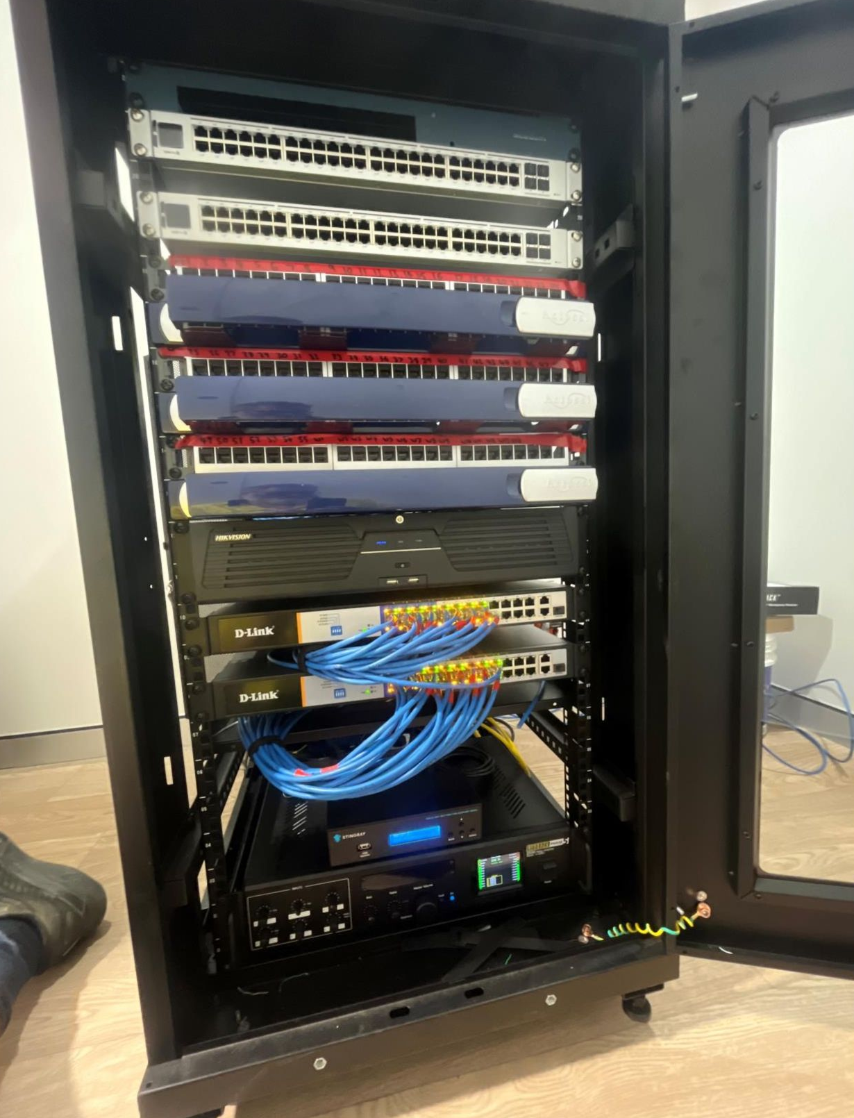 A server rack cabinet filled with network equipment and cabling, mostly blue and red, against a wall— Ray Smith Electrical in Korora, NSW