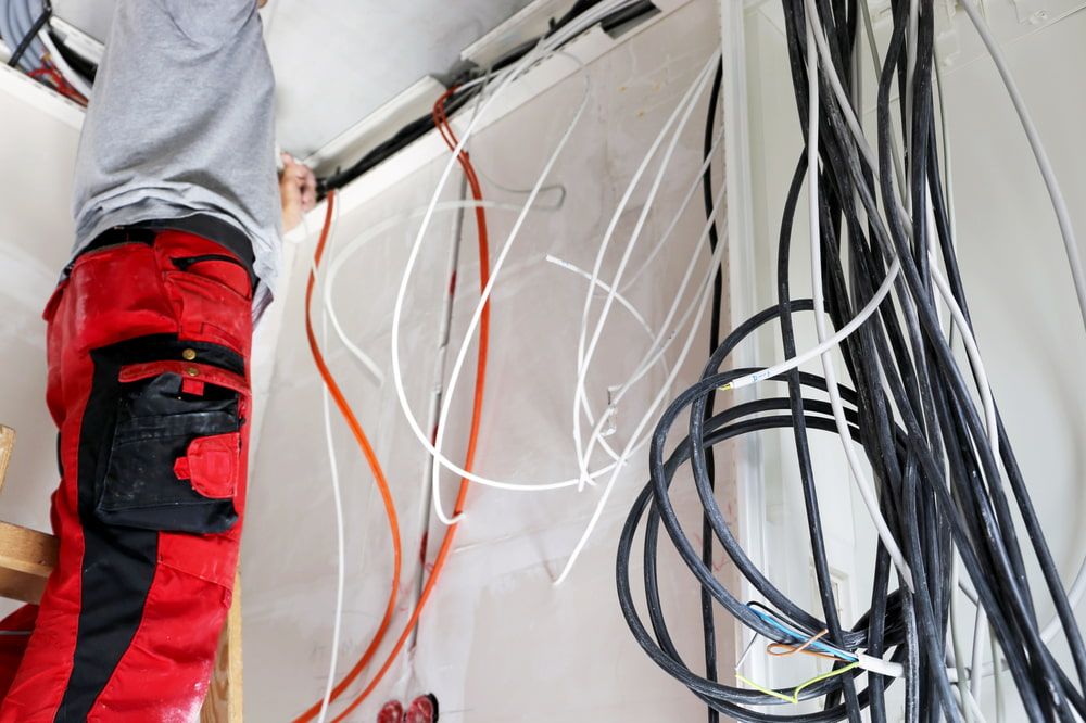 A Man in Red Pants is Working on a Wall With a Bunch of Wires Hanging From It — Ray Smith Electrical in Woolgoolga, NSW