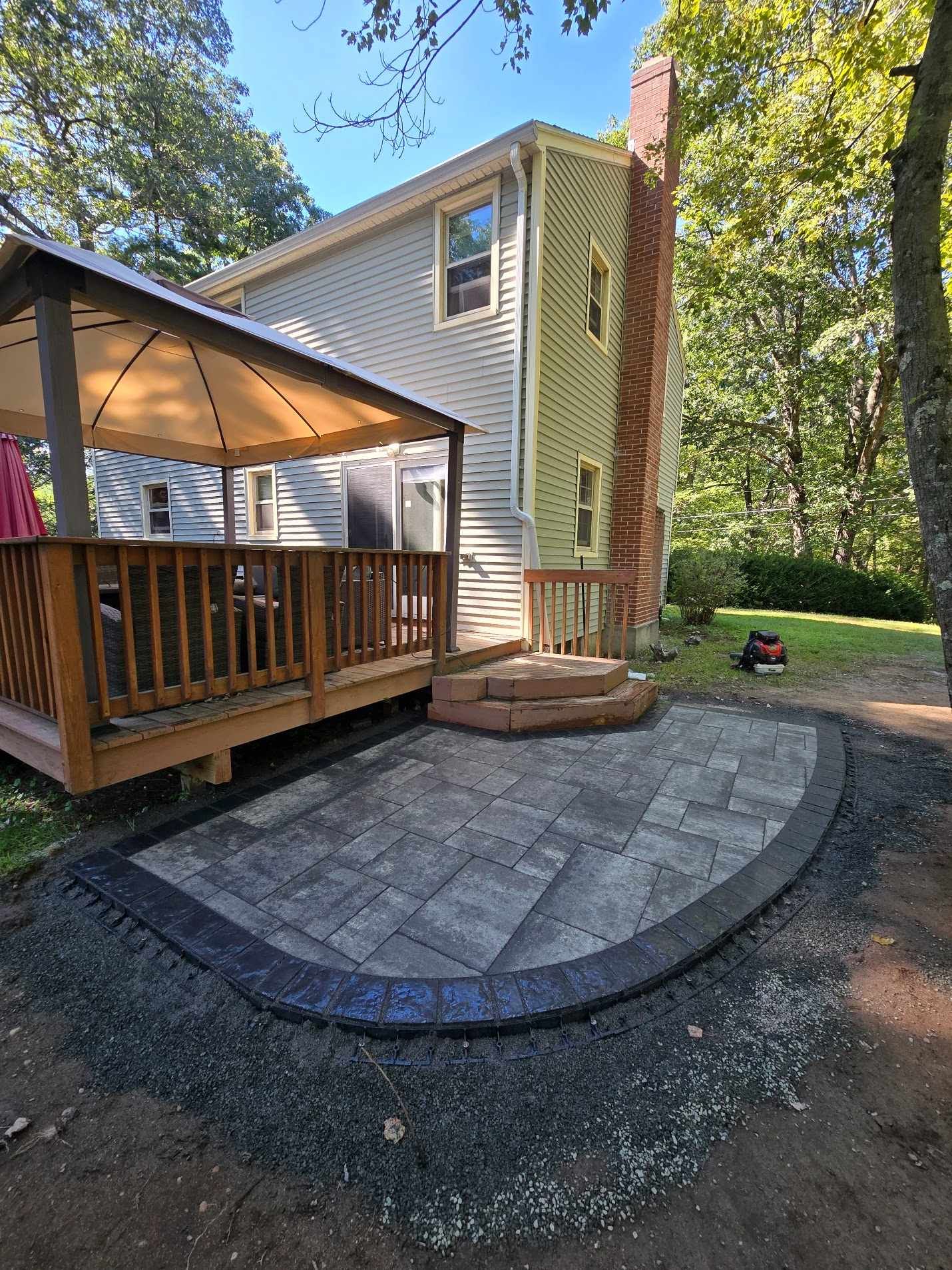 A backyard features a newly installed gray stone patio with a dark border, next to a wooden deck, gazebo, and house.