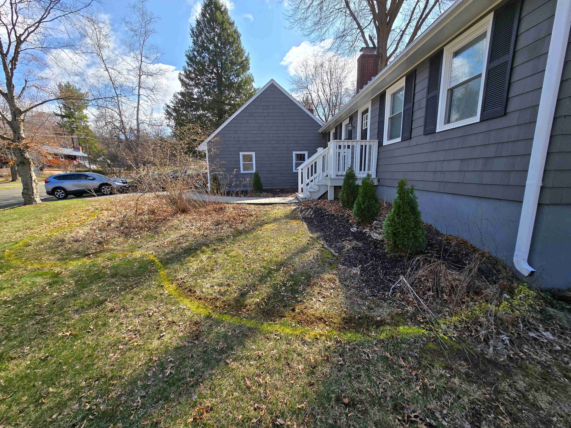 Gray house with a dark mulch flower bed marked by a yellow line, bordered by lawn and a small detached garage.
