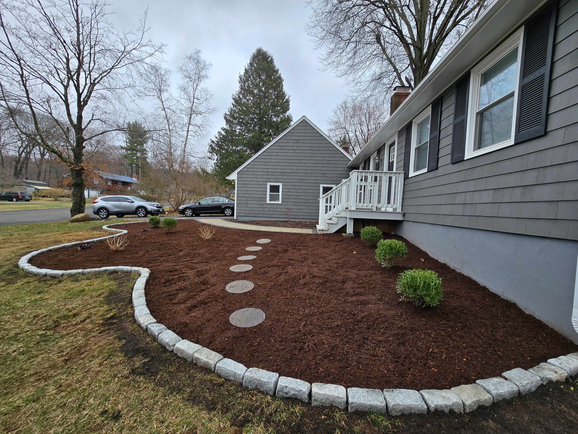 A gray house with new mulch, stone edging, and round stepping stones leading to a white deck on a cloudy day.