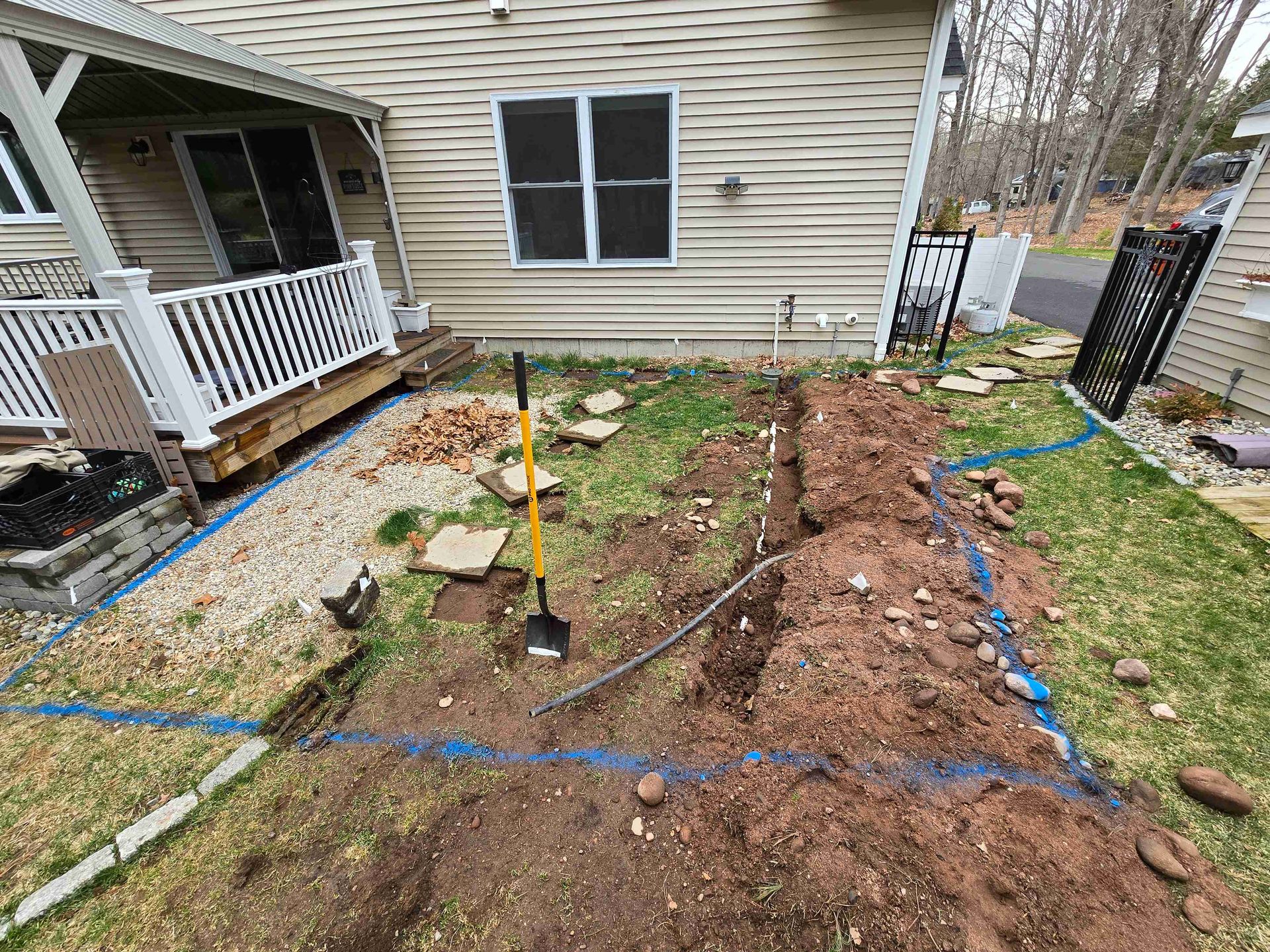A house exterior with a fenced deck and a dug-out trench marked with blue tape in the lawn for a landscaping project.