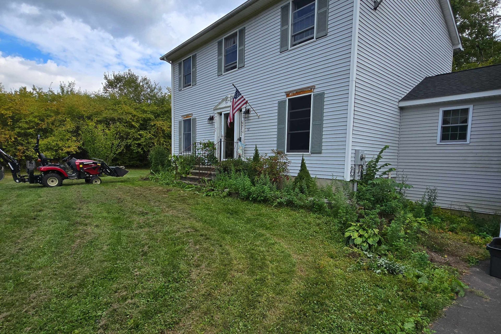 A white, two-story house with a lawn, American flag, and a red riding lawnmower parked on the left side.
