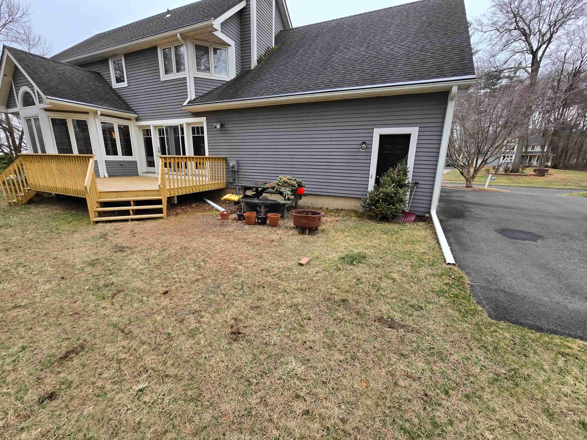 A gray two-story house with a wooden deck, a small lawn, and an adjacent asphalt driveway under a cloudy sky.