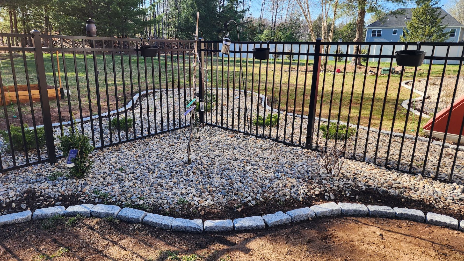 Black metal fence encloses a landscaped garden bed filled with gravel, small plants, and a young tree.