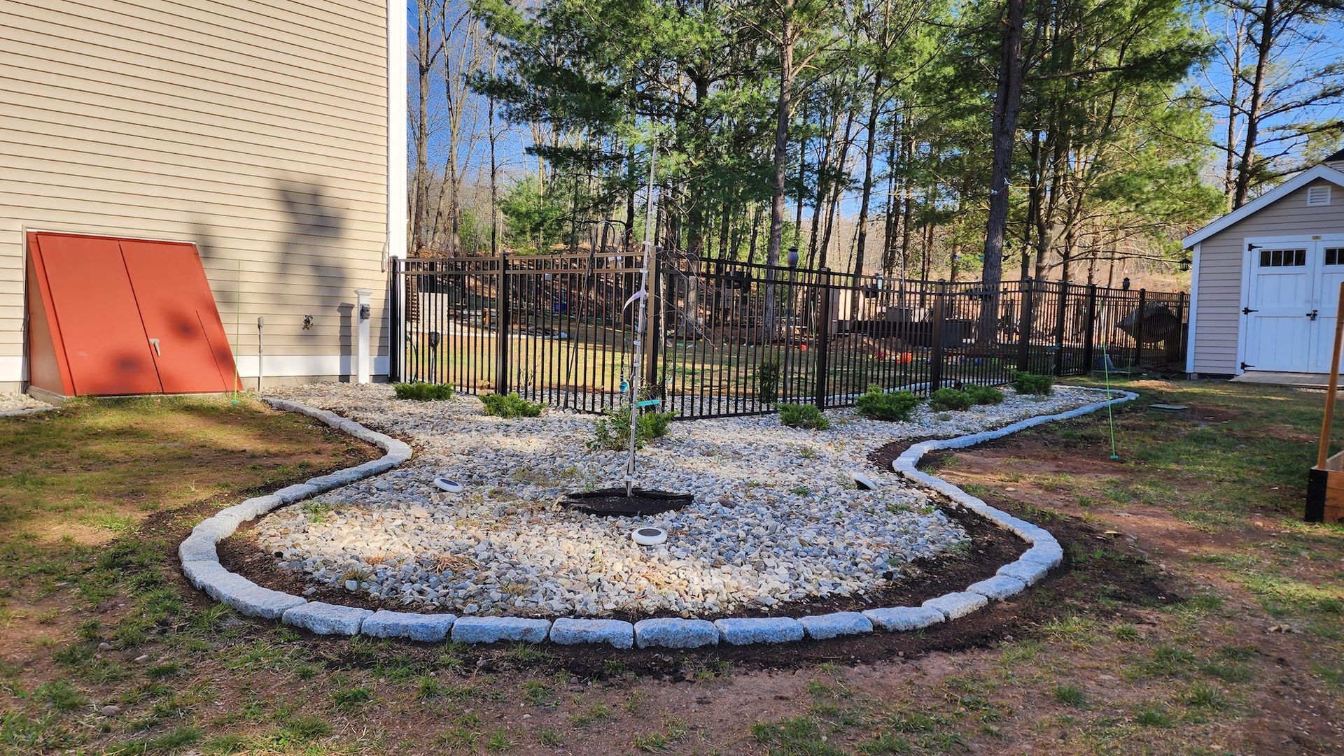 A rock garden bordered by gray stones in a yard, with a small shed and tall trees in the background.