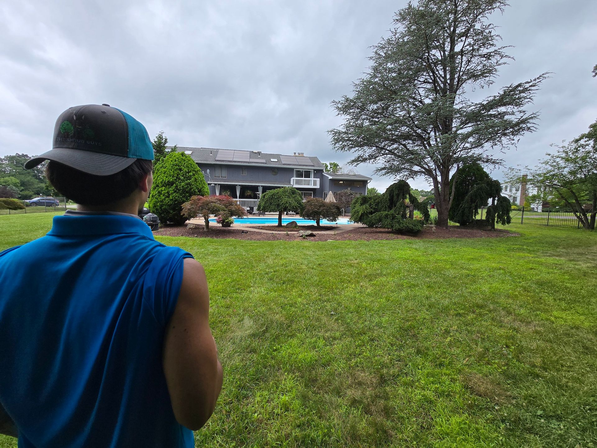 Person in blue shirt and cap looking at a backyard with a pool, trees, and a large house under a cloudy sky.
