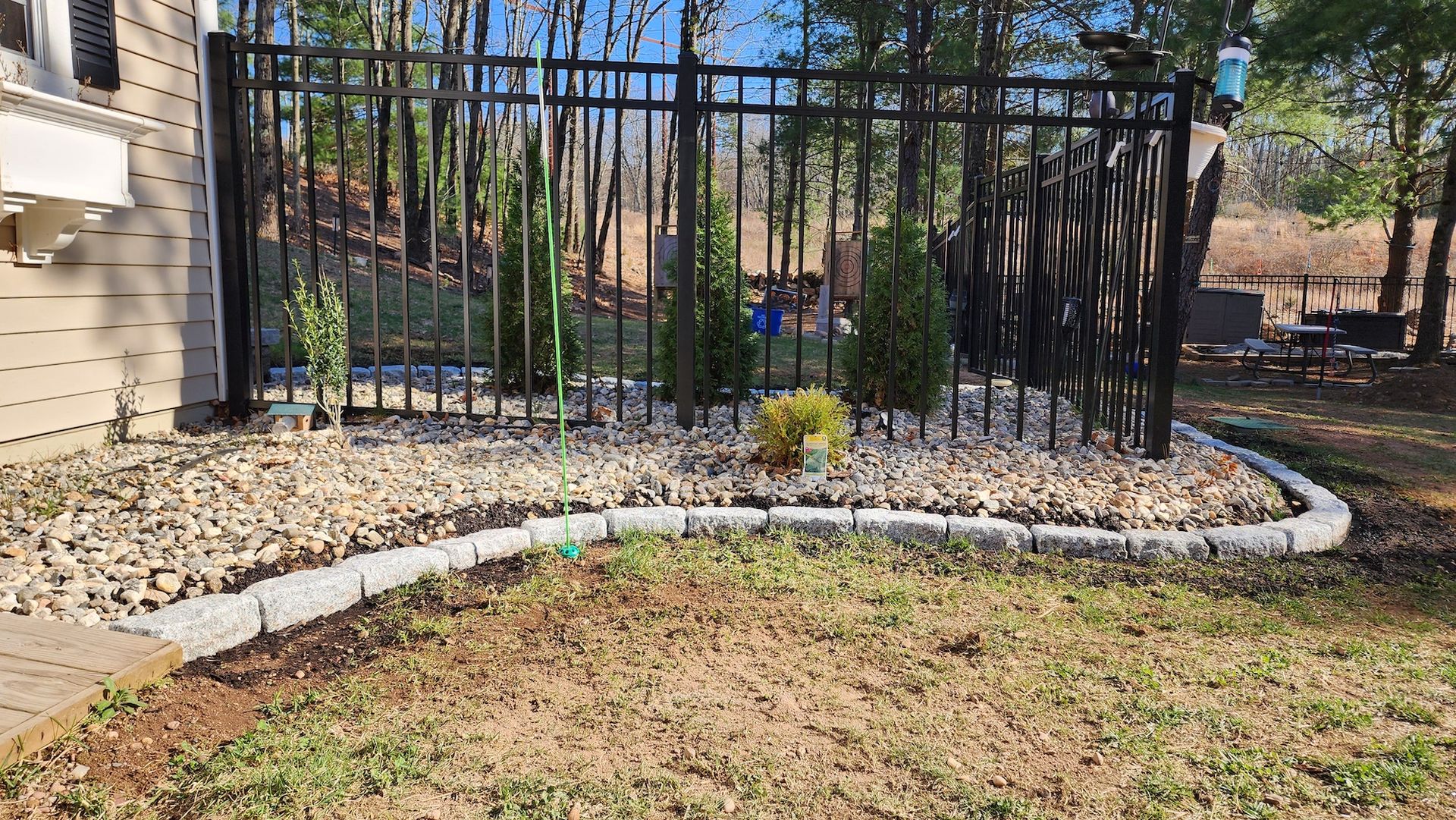 A black fence surrounds a rock-filled garden bed with small evergreen trees. A gray border separates the bed from the grass.