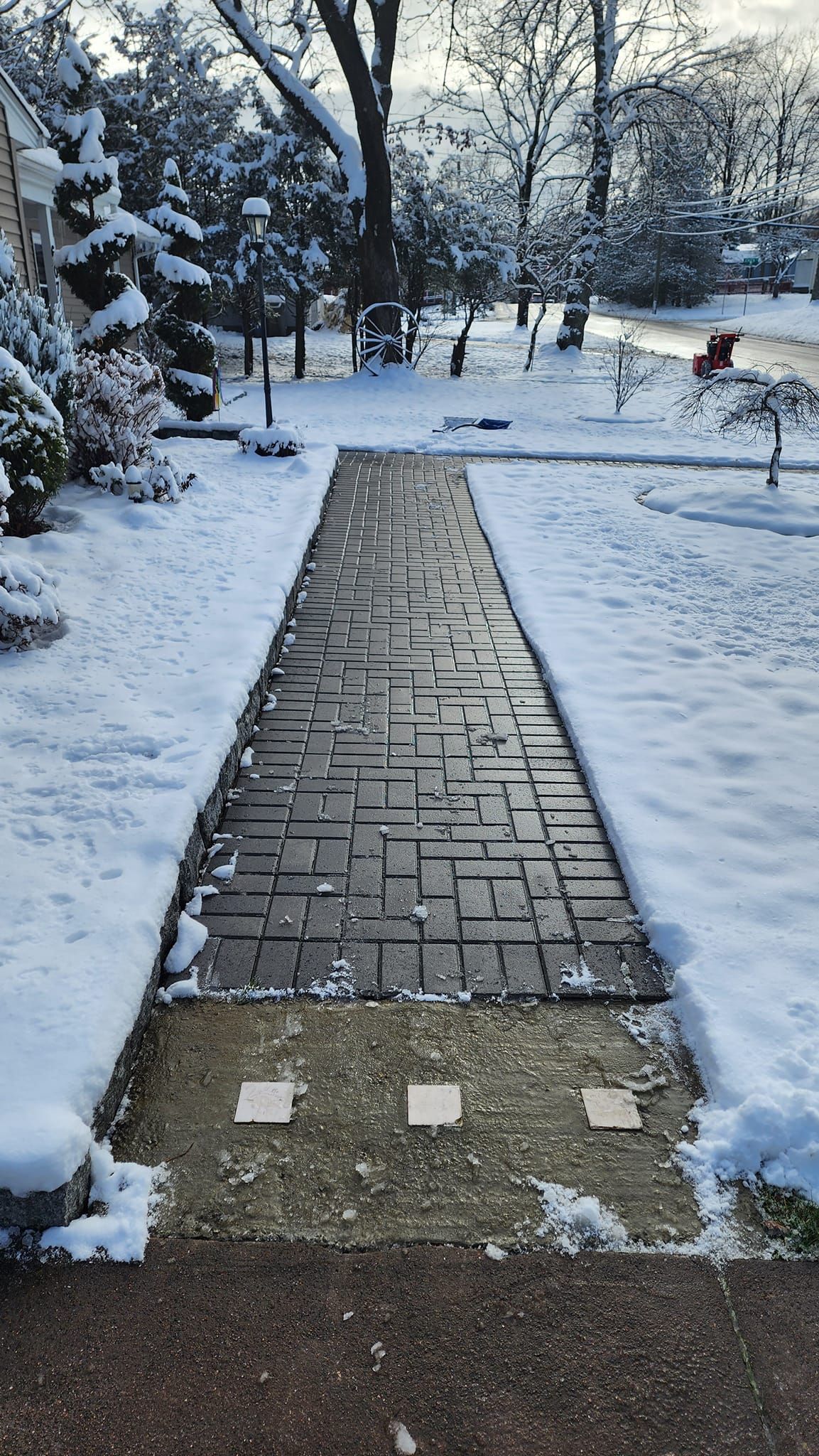 A snow-covered walkway of dark rectangular stones leads through a snowy park towards trees.