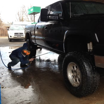 Man washing a large black truck at a car wash, using a hose. A white car is in the background.