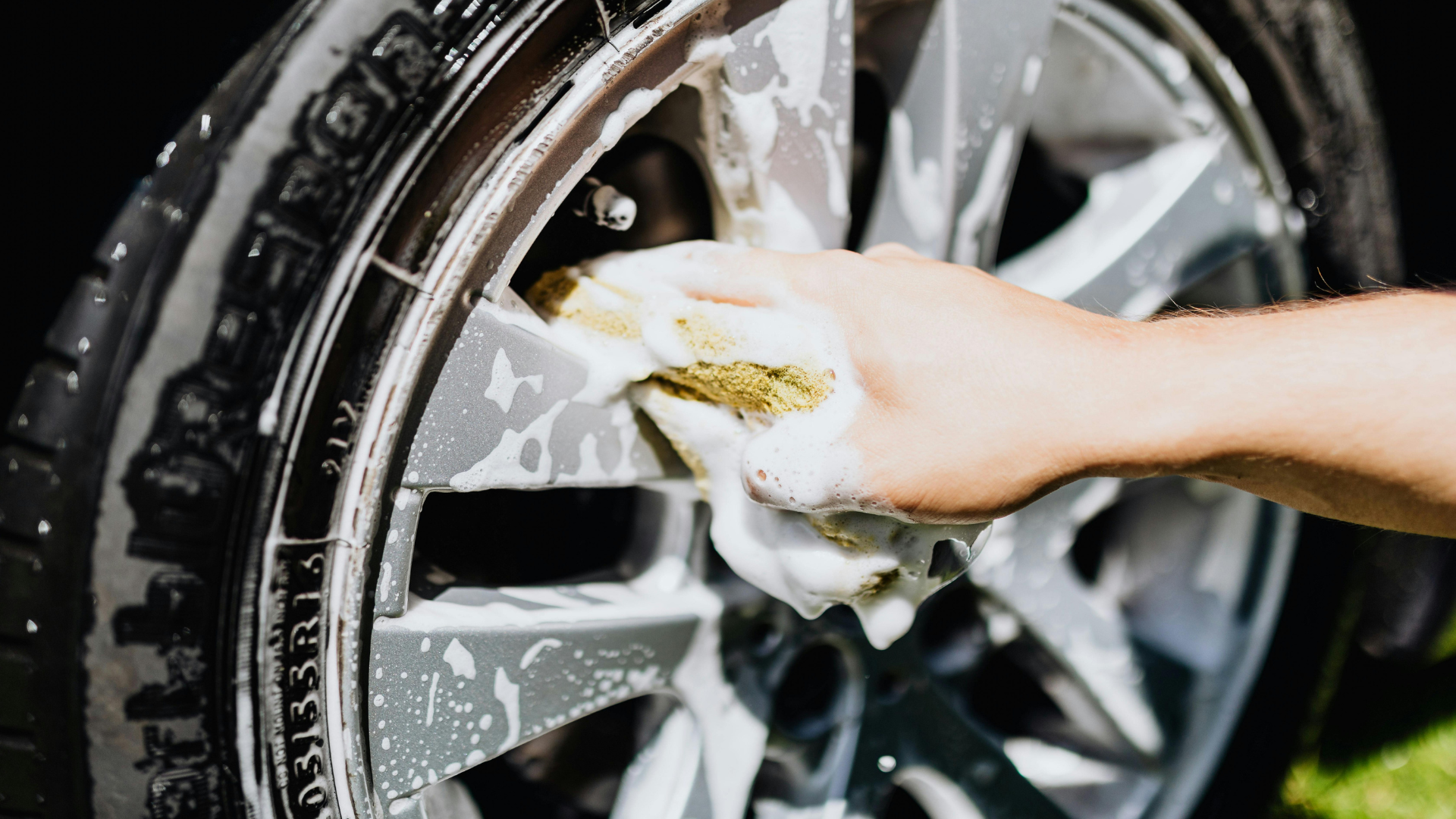 A person cleaning a car's silver wheel with a soapy cloth.