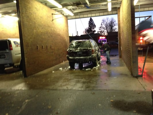 Car being washed at a carwash. A person is spraying soap on the black car, which is enclosed.
