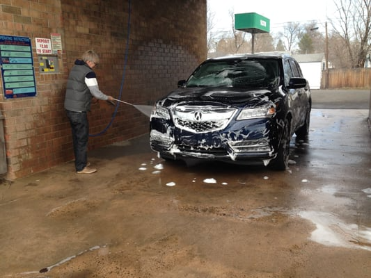 A man washing a dark blue Acura SUV at a car wash with soap and a water hose.