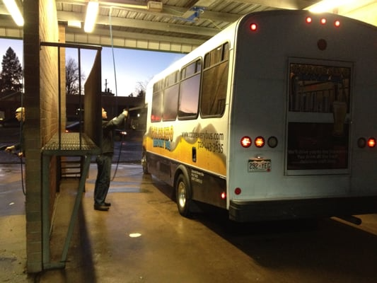 Person washing a bus at a car wash. Bus is white and yellow, person is holding a hose.