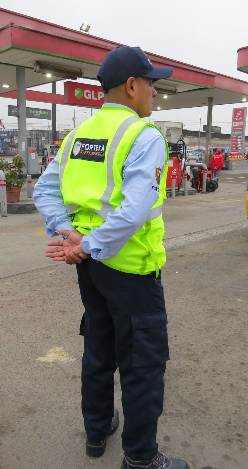 Un agente de tráfico con un chaleco amarillo neón está de pie con las manos detrás de la espalda en una gasolinera.