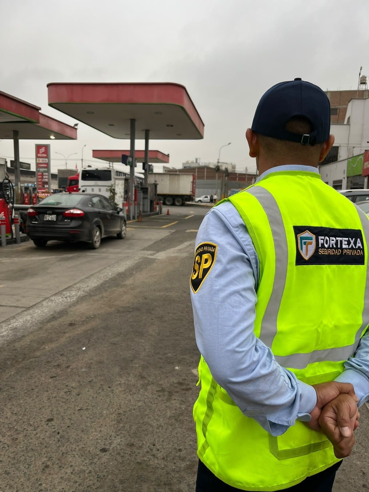 Un guardia de seguridad con chaleco reflectante vigila los coches en una gasolinera bajo un cielo nublado.