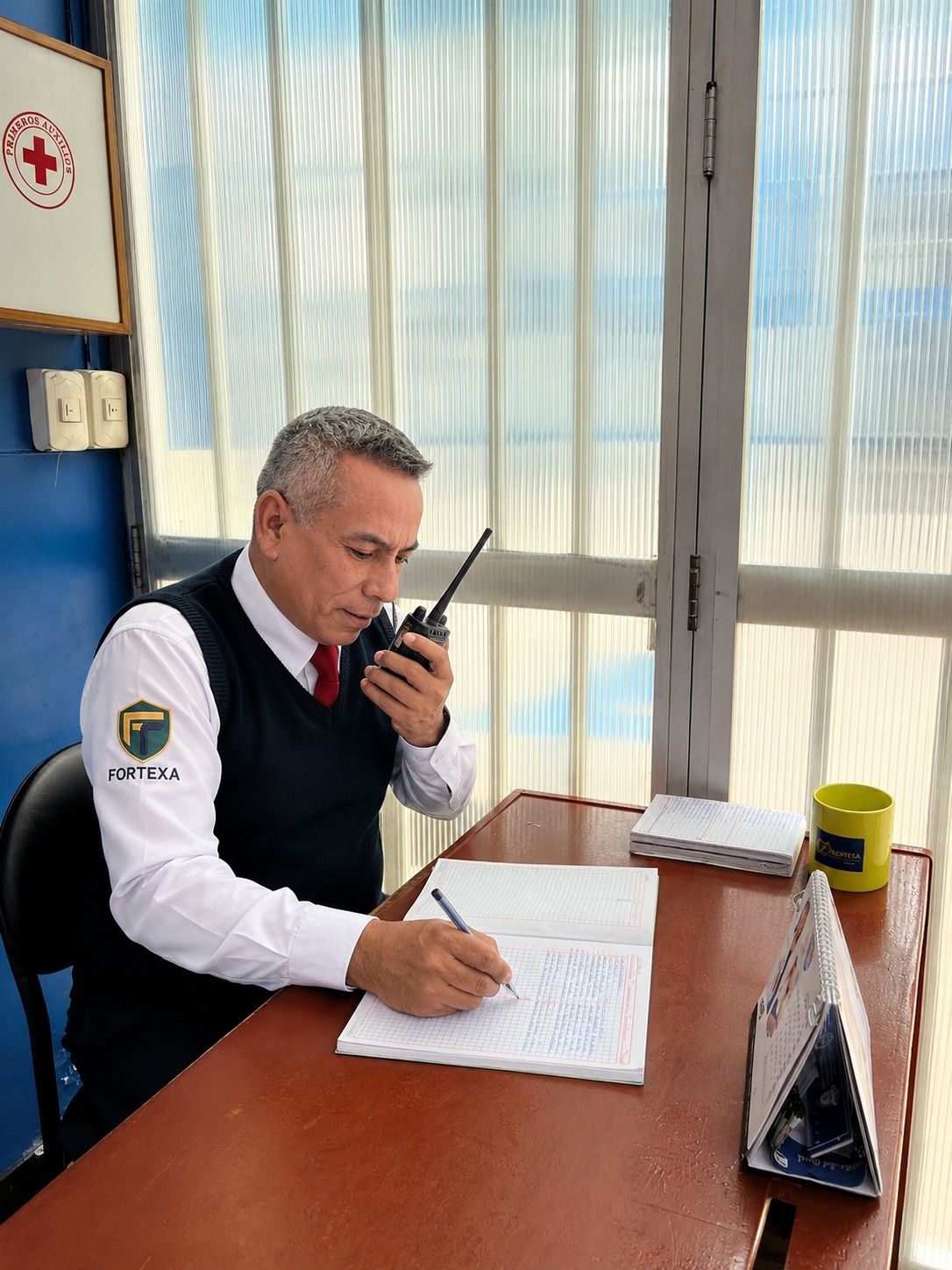 Un hombre con camisa blanca y chaleco oscuro habla por la radio mientras escribe en un escritorio