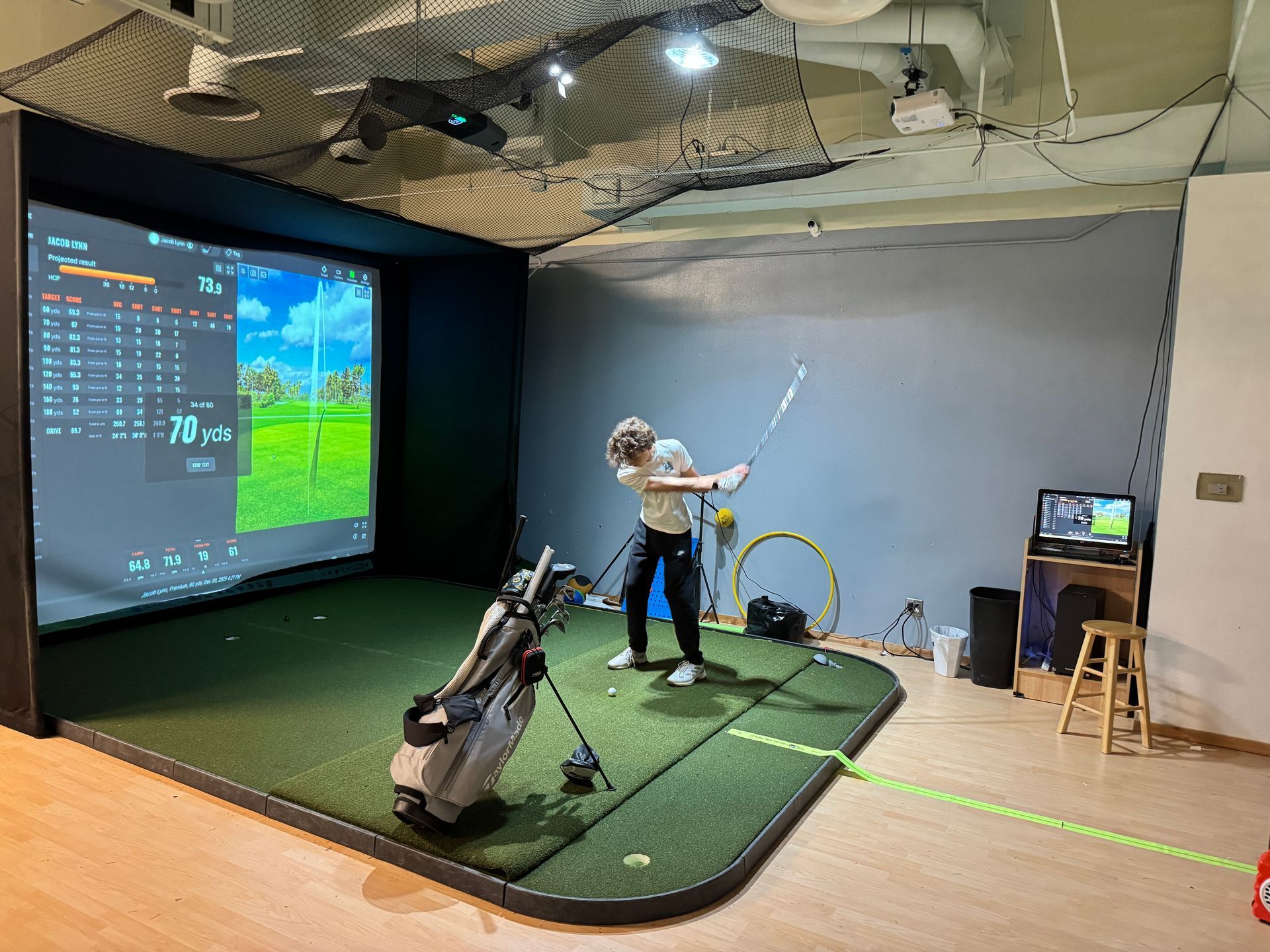 Indoor golf simulation room, man swinging club, two screens, artificial turf.