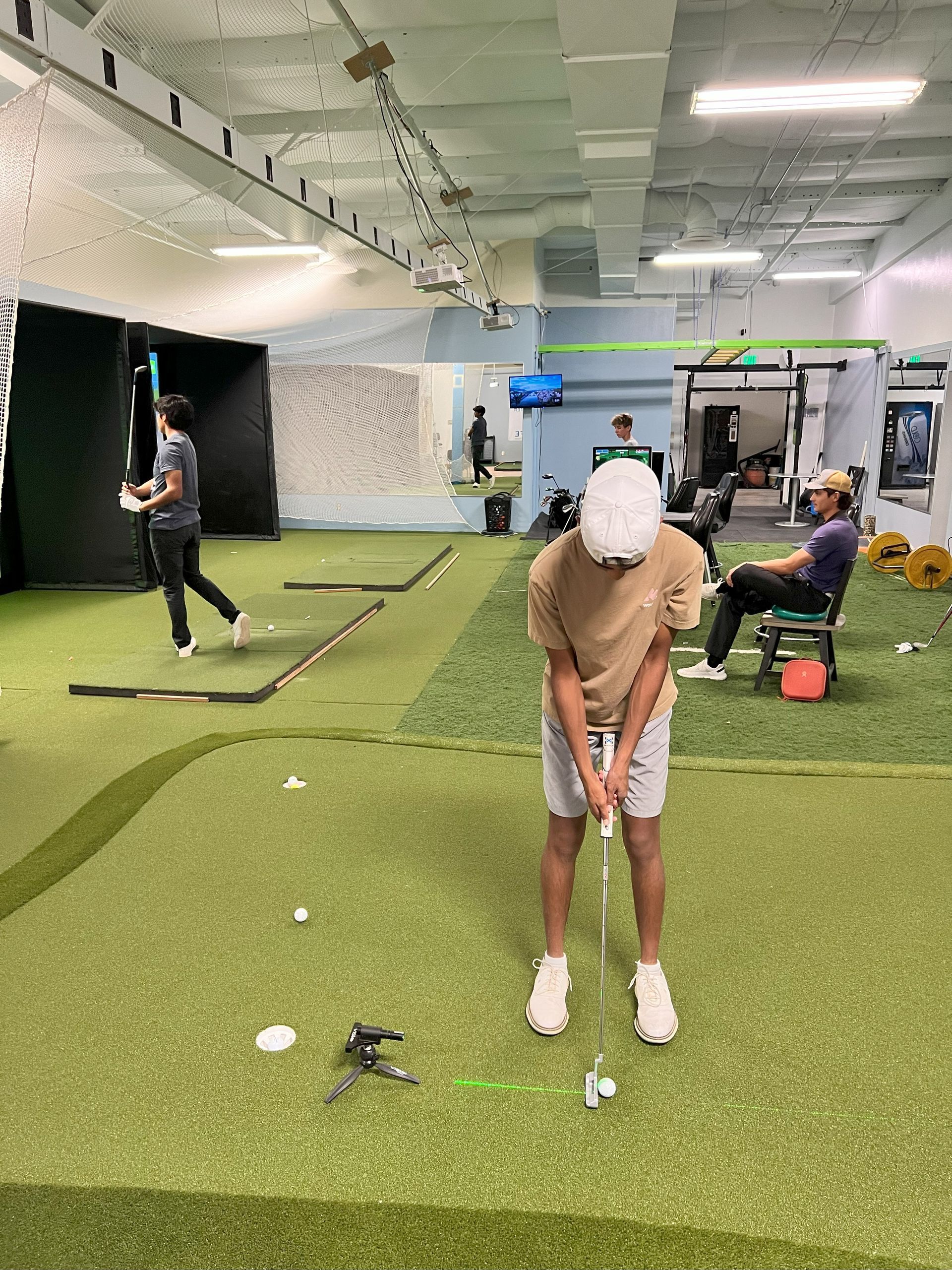 A person in a golf stance indoors, practicing putting. Others nearby, in a golf training area.